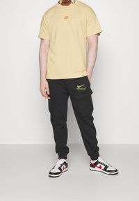 Nike t-shirt med rund hals i ljusgul med orange logo, kombinerad med svarta cargo-byxor med gul text, och svarta och vita sneakers.
