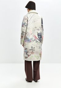 Persona con un cappotto chiaro con stampe astratte colorate di natura e animali, pantaloni larghi marroni e un cappello marrone scuro, in piedi.