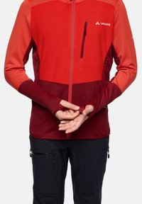 Rote und kastanienbraune Zip-Jacke mit strukturiertem oberen Teil, eng anliegenden Ärmeln mit Daumenlöchern und einer Brusttasche. Getragen über schwarzen Hosen.