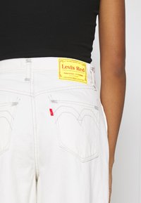 Bílé denimové džíny se žlutou červenou visačkou Levi's na zadní straně, s charakteristickou červenou tabulkou a zakřiveným prošíváním na kapsách.