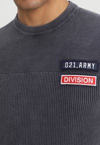 Grauer, strukturiert gestrickter Pullover mit Rundhalsausschnitt und zwei gestickten Aufnähern: "021.ARMY" in Dunkelblau und "DIVISION" in Rot auf Weiß.