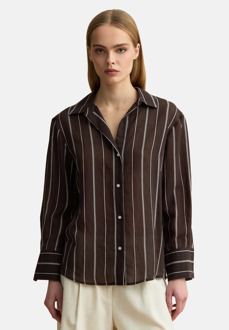Frau trägt ein dunkelbraunes langärmeliges Hemd mit vertikalen weißen Streifen und beigen Hosen vor einem schlichten Hintergrund.