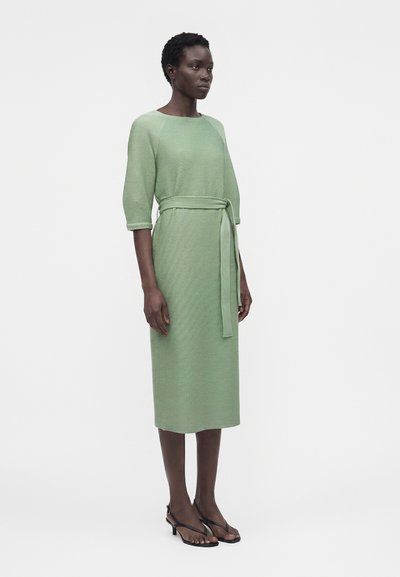 Γυναίκα που φοράει ένα ανοιχτοπράσινο, υφασμάτινο midi φόρεμα με ζώνη στη μέση και μανίκια τριών τετάρτων, συνδυασμένο με μαύρα σανδάλια με τακούνι.
