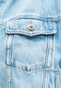 Poche d'une veste en jean bleu clair avec rabat sécurisé par un bouton argenté gravé du nom de la marque "DRYKORN".