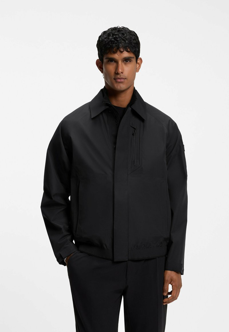 Young man with curly hair wearing a black zip-up jacket with collar and black pants, standing against a plain white background.