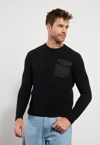 Pull noir côtelé avec un col rond et des manches longues. Il présente une poche poitrine noire mate avec une fermeture à pression. Associé à un jean bleu clair.