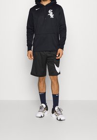 Homme portant un sweat à capuche noir des Chicago White Sox, des shorts noirs Nike, des chaussettes Nike bleu marine et des chaussures de sport blanches, debout devant un fond uni.