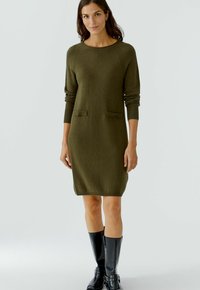 Olivgrünes Strickkleid mit langen Ärmeln, rundem Halsausschnitt und zwei Vorder-Taschen. Gerippte Bündchen und Saum, kombiniert mit schwarzen kniehohen Stiefeln.