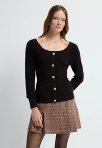 Zwarte geribde cardigan met lange mouwen, een vierkante halslijn en vijf gouden knopen. Gecombineerd met een gestructureerde bruine ruitrokken.