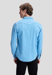 Man met lichtbruin haar die een lichtblauw denim overhemd met lange mouwen draagt, van achteren gezien tegen een effen grijze achtergrond.