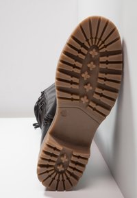 Sole černé boty s tlustou, vzorovanou hnědou gumovou podrážkou, která má křížové a liniové designy, odpočívající na bílé ploše.