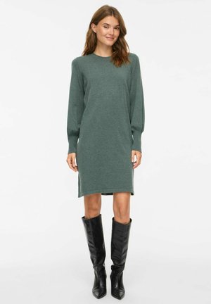 Femme debout portant une robe en maille verte à manches longues et des bottes noires à bout pointu montant jusqu'aux genoux sur un fond blanc uni.