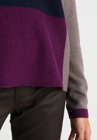 Sweter z dzianiny w wielokolorowym wzorze z ciemnoniebieskim, fioletowym i szarym. Materiał jest miękki, z lekko szczotkowaną strukturą.