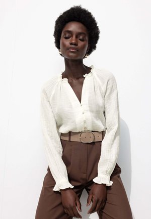 Blouse blanche avec col et poignets froncés, fermeture à boutons à l'avant, associée à un pantalon marron taille haute et une ceinture beige avec une boucle surdimensionnée.