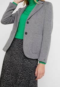 Blazer grigio con due bottoni e rever a incavo, abbinato a un dolcevita verde e a una gonna stampa leopardo nera e grigia.