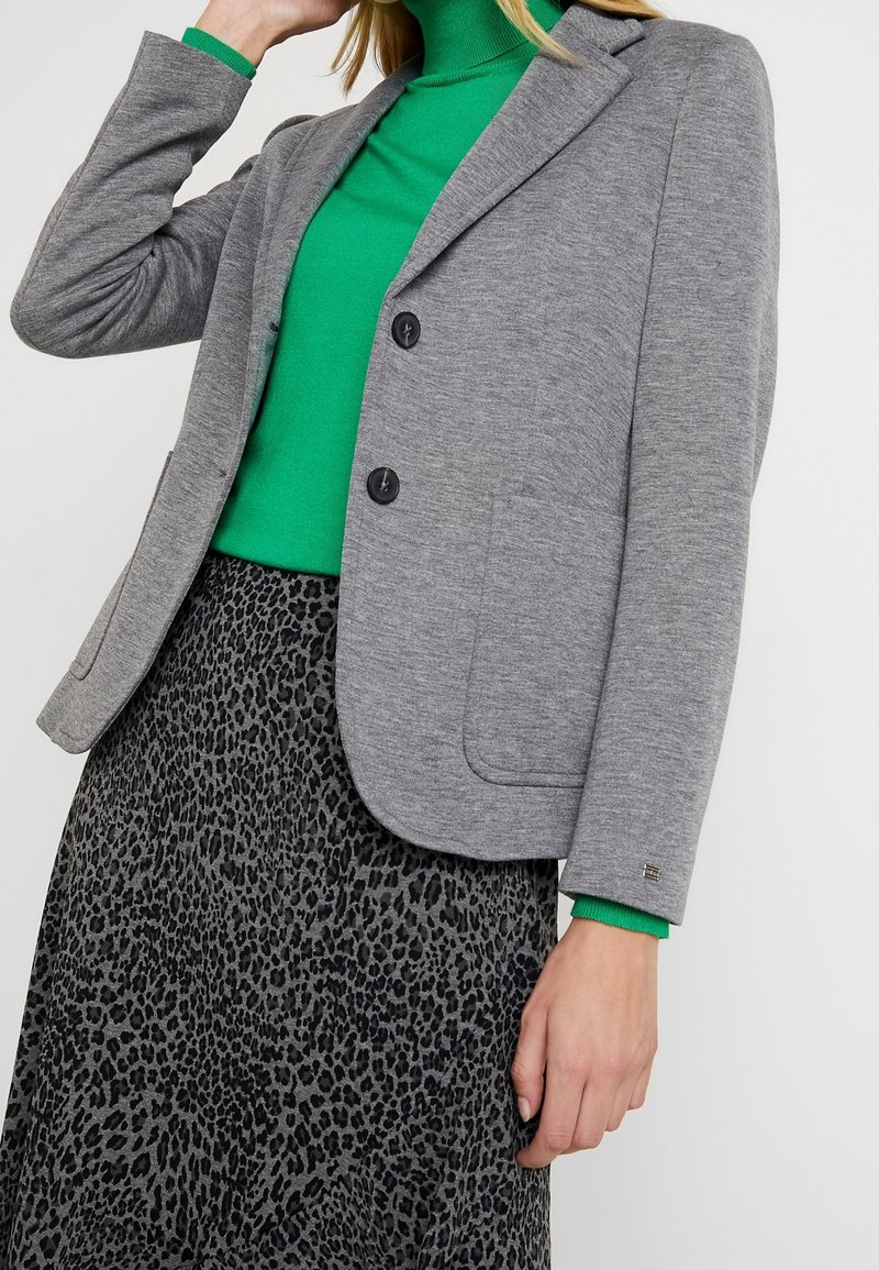 Blazer grigio con due bottoni e rever a incavo, abbinato a un dolcevita verde e a una gonna stampa leopardo nera e grigia.