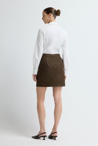 Witte blouse met lange mouwen gecombineerd met een bruine, gestructureerde minirok. Zwarte slingback hakken met een gebogen hakontwerp. Effen achtergrond.