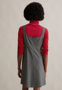 Grau gestreiftes Kleid mit tailliertem Oberteil und rundem Rückenausschnitt, über einem figurbetonten roten Rollkragenpullover getragen. Glattes Material, ärmelloses Design.