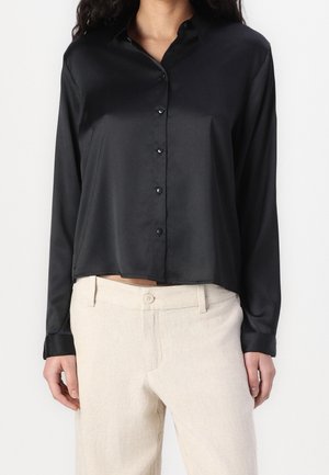 Black satin button-up shirt with long sleeves and a classic collar. Features a cropped hem. Paired with light beige trousers for contrast.