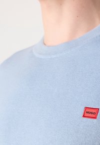 Gros plan d'une personne portant un pull tricoté bleu clair avec un petit patch rectangulaire rouge portant l'inscription « HUGO » sur la poitrine supérieure.