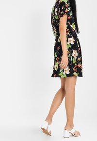 Kvinna i en svart blommig klänning och vita klacksandaler, stående med ena foten något lyft mot en enkel vit bakgrund.