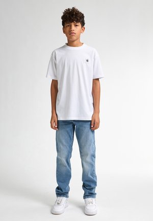 Witte t-shirt met korte mouwen en een klein logo op de linkerborst, gecombineerd met lichtblauwe jeans en witte sneakers, staand op een neutrale achtergrond.