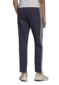 Pantaloni della tuta blu navy con strisce laterali bianche, vestibilità tapered, vita elasticizzata e tasca posteriore. Indossati con scarpe da ginnastica di colore chiaro.