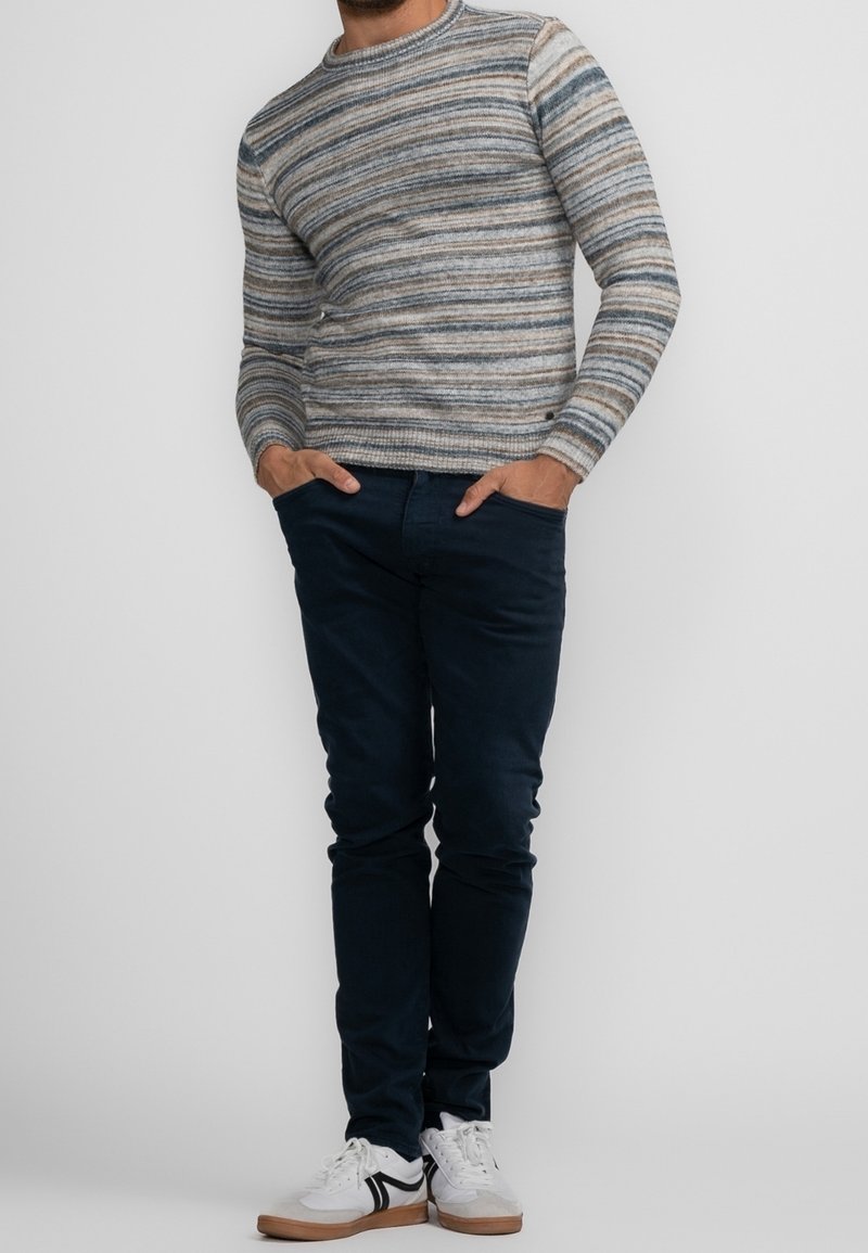 Homme portant un pull rayé à manches longues, pantalon bleu foncé, baskets blanches avec des accents noirs, debout les mains dans les poches devant un fond uni.