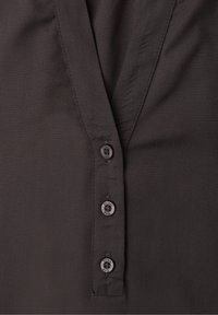 Gros plan sur un tissu de chemise marron foncé avec une patte verticale présentant trois petits boutons ronds étiquetés "Street One."
