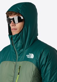 Groene pufferjack met capuchon, voorzien van een lichter groen paneel, wit logo en een ritsborstzak met een felgroene rits.