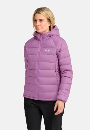 Vrouw draagt een lichtpaarse gewatteerde jas met capuchon en zwarte broek, staand tegen een effen lichte achtergrond.