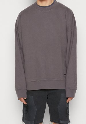 Person trägt einen oversized, langärmeligen grauen Pullover und schwarze, ausgewaschene Jeansshorts und steht vor einem schlichten weißen Hintergrund.