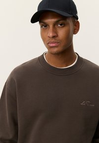 Καφέ φούτερ με ριμπ λαιμόκοψη, διακοσμημένο με ανοιχτό καφέ κεντημένο κείμενο. Μαύρο καπέλο συμπληρώνει την εμφάνιση, με τα δύο προϊόντα εμφανισμένα από κοντά.