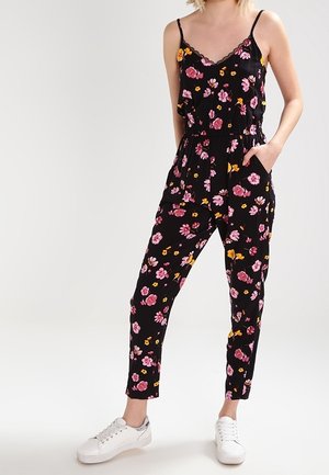 Vrouw draagt zwarte bloemenmouwloze jumpsuit met roze en gele bloemen en witte sneakers, staand met één hand in de zak.