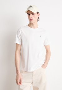 Hvit bomulls T-skjorte med rund hals, med en liten brodert logo. Kombinert med lyse beige bukser og en kremfarget baseballcaps.