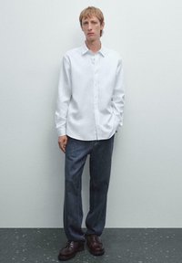 Weißes langärmliges Button-Up-Hemd, kombiniert mit locker sitzenden dunklen Jeans und dunklen Schnürschuhen, vor einem schlichten hellen Hintergrund.
