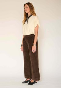 Haut crème à manches courtes, pantalons en velours côtelé marron à jambes larges, chaussures noires en cuir à enfiler avec des accents cloutés, et une écharpe noire nouée autour du cou.