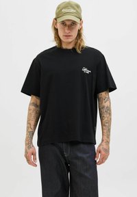 Camiseta de algodón negra con mangas cortas, que presenta un pequeño logo blanco en el lado izquierdo del pecho. Combinada con jeans de mezclilla oscura, que muestran un ajuste relajado.