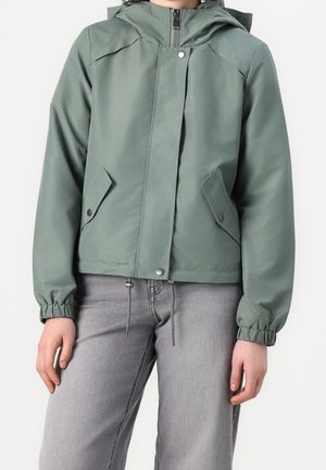 Person iført salviegrøn lynlåstrøje med trykknaplommer og elastiske manchetter, kombineret med lysegrå jeans.