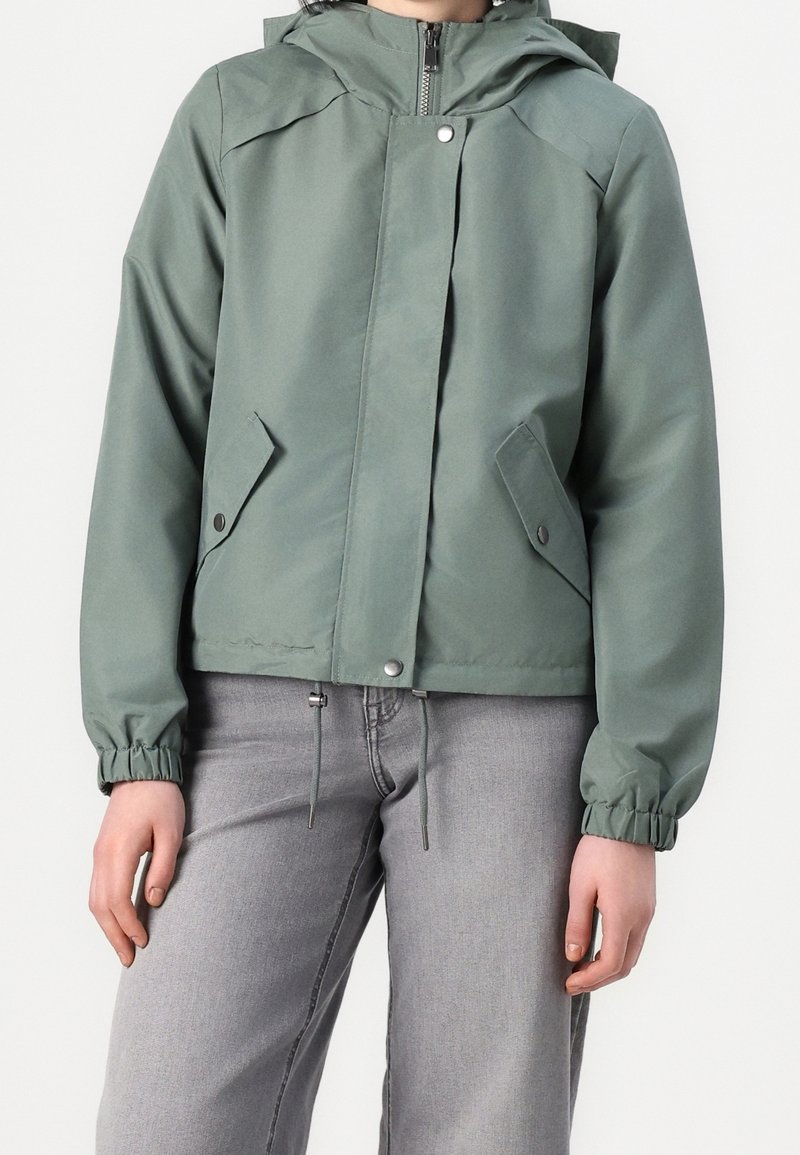 Personne portant une veste zippée vert sauge avec poches à boutons-pression et poignets élastiques, associée à un jean gris clair.