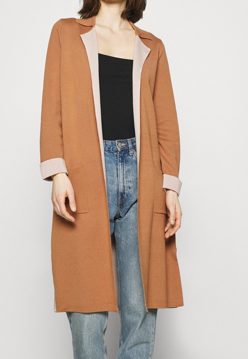 Femme portant un long manteau marron avec des poignets beiges, un haut noir et un jean taille haute bleu clair sur un fond blanc uni.