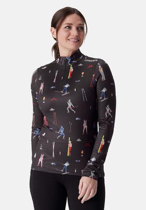 Femme portant un t-shirt noir à manches longues sur le thème du ski avec des imprimés colorés de skieurs et d'équipements de ski, souriante et regardant sur le côté.
