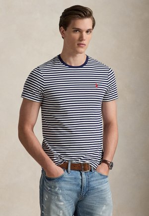 Jeune homme portant un t-shirt rayé bleu marine et blanc, un jean bleu avec une ceinture marron et une montre-bracelet, debout les mains dans les poches.