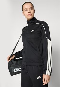 Fato de treino preto da Adidas com riscas brancas nas mangas, casaco com fecho éclair, combinado com uma bolsa preta com o logótipo da Adidas. Textura lisa.
