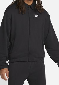 Svart zip-up sportjacka i mjukt tyg, med krage, elastiska ärmslut och en liten vit Nike-logga på bröstet.