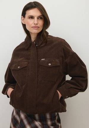 Vrouw met bruin haar die een bruine corduroy jas met voorzakken en een geruite rok draagt, kijkt opzij met handen in de zakken.