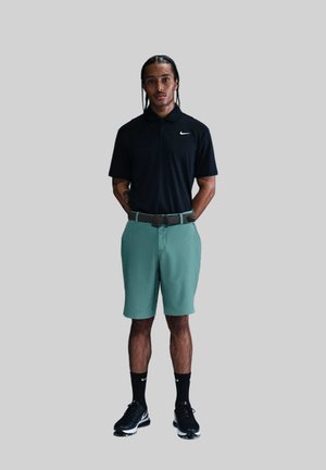 Svart pikétröja med vit Nike-logotyp, gröna shorts, svarta skor och svarta strumpor. Man står med händerna bakom ryggen. Enkel bakgrund.