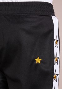 Schwarze Sporthose mit elastischem Bund, seitlichen Streifen und gelben gestickten Sternen entlang des Beins. Glatte Stofftextur.