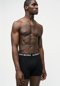 Μυώδης άντρας που στέκεται, φοράει μαύρα μποξεράκια Björn Borg, κοιτάζει αριστερά του μπροστά σε ένα απλό ανοιχτό φόντο.