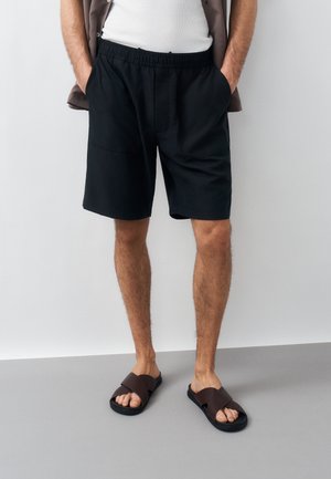 Man draagt zwarte elastische tailleband short, wit hemdje, bruine sandalen met open tenen, staat met handen in de zakken tegen een effen achtergrond.